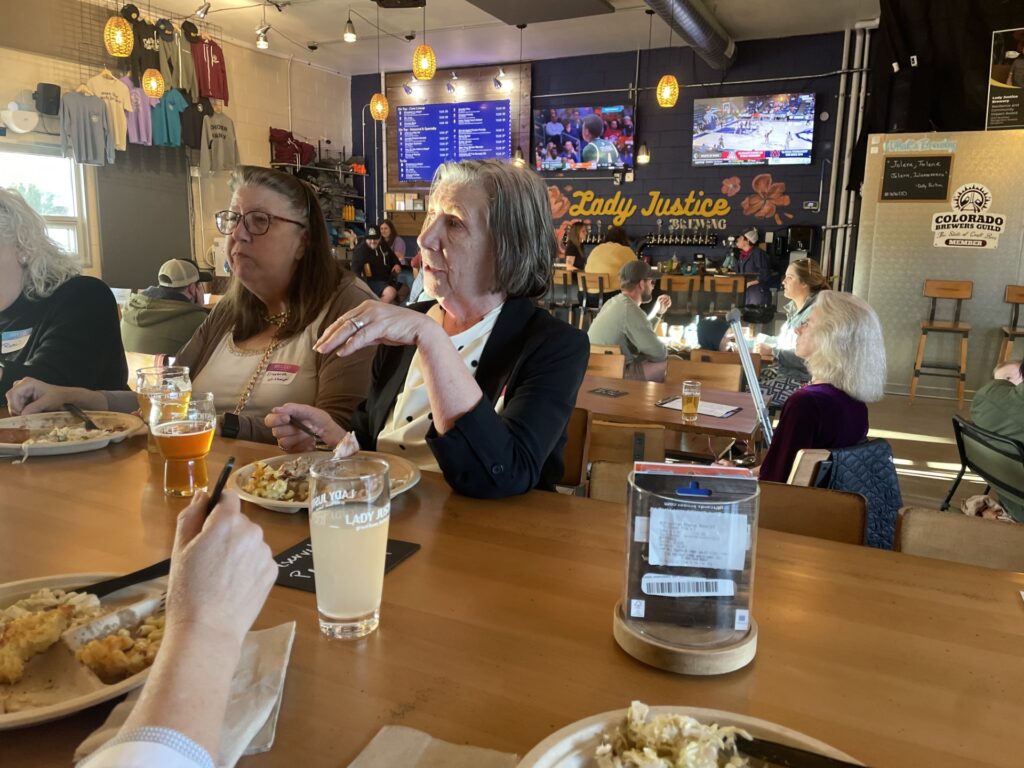 Women sitting at a tall table eating and drinking beer in a brewery tap room with the mural of Lady Justice Brewing on the back wall